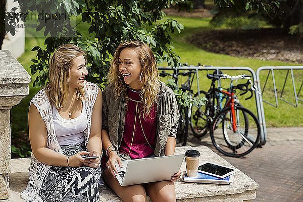 Zwei Studentinnen lachen und unterhalten sich  während sie draußen auf dem Campus ihre Technik benutzen  Edmonton  Alberta  Kanada