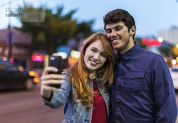 Ein junges Paar steht in der Abenddämmerung an einer Straße in der Stadt und macht ein Selbstporträt mit einem Smartphone  Edmonton  Alberta  Kanada