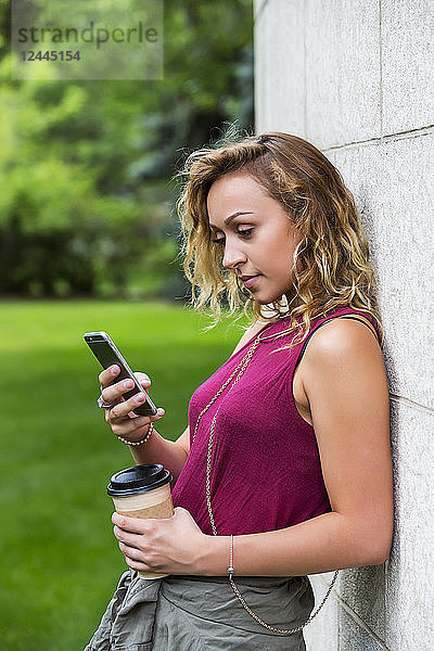 Eine hübsche junge Studentin lehnt an einer Wand auf dem Campus und liest Texte auf ihrem Smartphone  Edmonton  Alberta  Kanada