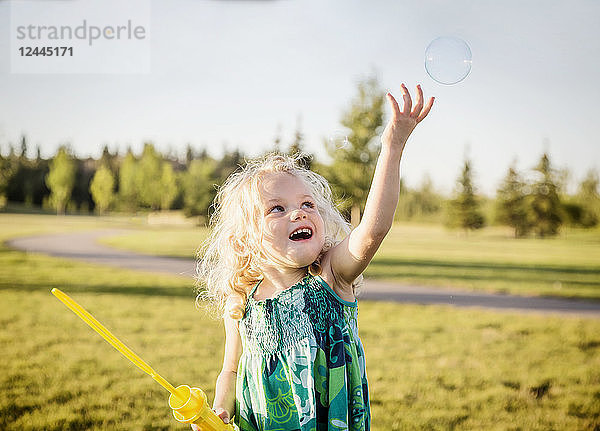 Ein süßes junges Mädchen mit blondem Haar  das eine Seifenblase aufbläst und versucht  sie in einem Park an einem warmen Herbsttag zu fangen  Fichte