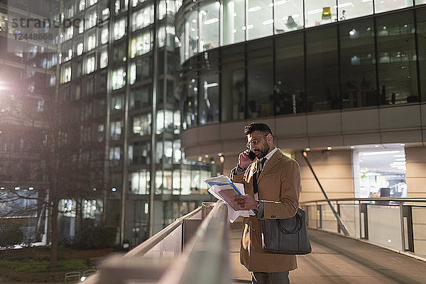 Businessman talking on smart phone and reading paperwork on urban pedestrian bridge at night