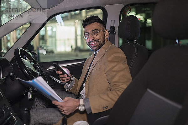 Portrait smiling businessman with smart phone reading paperwork in car at night