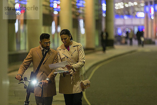 Business people with bicycle and paperwork walking on urban street at night
