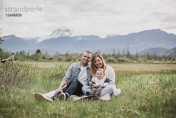 Portrait smiling parents and baby son sitting in rural field with mountains in background