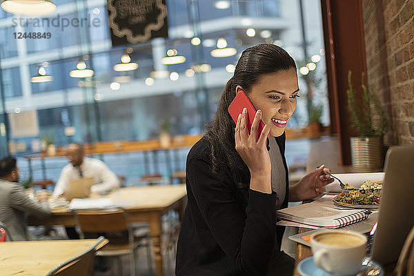 Smiling businesswoman talking on smart phone and working at laptop in cafe