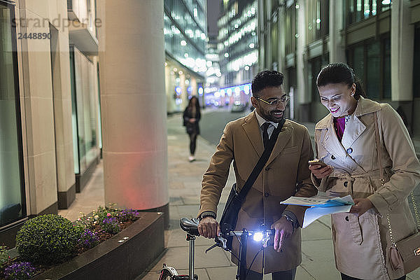 Business people with smart phone and bicycle reviewing paperwork on urban street at night
