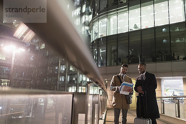 Businessmen with suitcase discussing paperwork on urban pedestrian bridge at night
