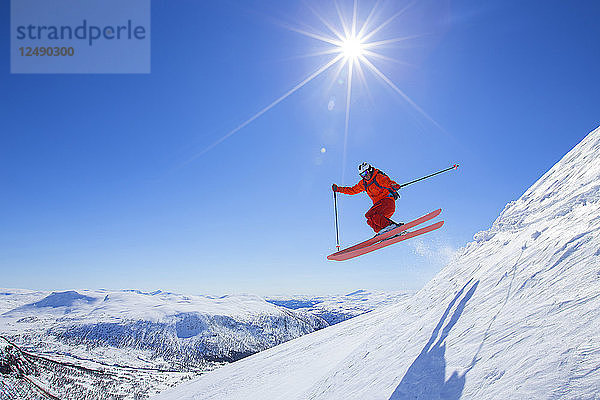 Ein männlicher Freerider in einem roten Anzug springt von einem Schneekamm. Die Sonne scheint  der Himmel ist blau. Myrkdalen in der norwegischen Region Flord ist ein geheimes Pulverschnee-Paradies. Das Skigebiet liegt in der Nähe des Voss-Gebietes und zählt einen jährlichen Schneefall von bis zu 10 Metern Schnee.