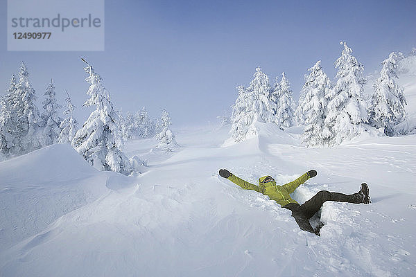 Ein Mann liegt glücklich im Tiefschnee auf einem alpinen Plateau  umgeben von schneebedeckten Fichten  und blickt in den Himmel. Er ist mit einer grünen wasserdichten Jacke  einer wasserdichten Hose  Handschuhen  Gamaschen und Winter-Trekkingstiefeln bekleidet.