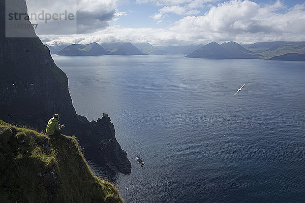 Männlicher Wanderer sitzt auf einem Felsvorsprung über dem Meer und blickt auf die fernen Berge  während eine Möwe vorbeifliegt.