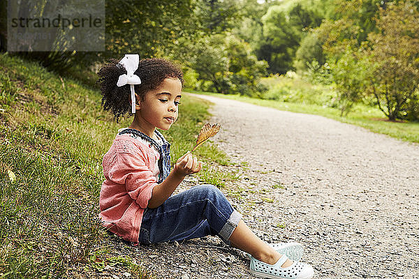 Ein kleines Mädchen sitzt auf der Seite der Straße und hält ein Herbstblatt