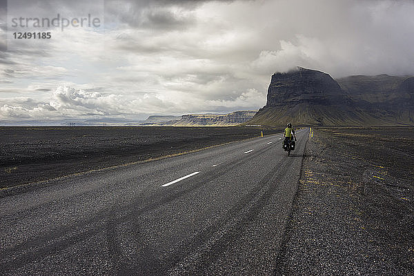 Frau fährt mit dem Tourenrad auf einer geraden Straße in Richtung eines Berges in einer vulkanischen Landschaft.