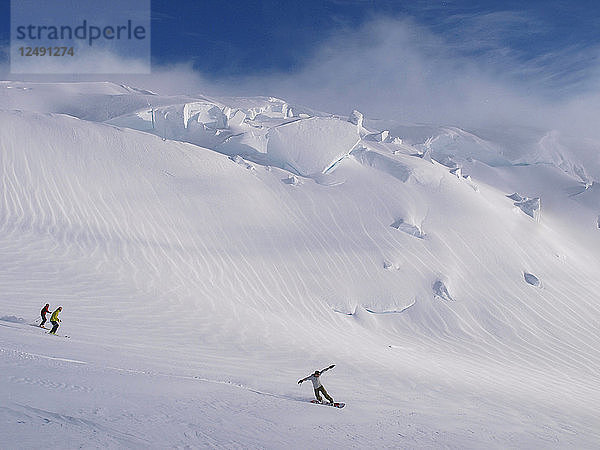 Bergsteiger fahren Ski und Snowboard im Abendlicht  in der Nähe des 12.000-Fuß-Camps auf einem Gletscher am Mount McKinley. Um sich zu akklimatisieren  fahren viele Alpinisten gerne ein paar Abfahrten in der Nähe des Lagers. Der Mount McKinley oder Denali ist der höchste Berg Nordamerikas mit einer Gipfelhöhe von 6.168 m (20.237 Fuß) über dem Meeresspiegel. Mit einer Höhe von rund 5.500 m (18.000 Fuß) ist er der höchste Berg  der vollständig über dem Meeresspiegel liegt. Gemessen an der topografischen Ausdehnung ist er nach dem Mount Everest und dem Aconcagua der drittgrößte Berg. Der McKinley befindet sich in der Alaska Range im Inneren des US-Bundesstaates Alaska und ist das Herzstück des Denali National Park and Preserve.