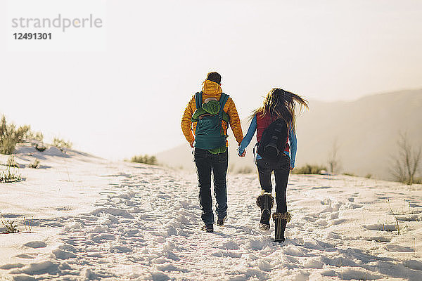 Ein junges Paar hält sich an den Händen und wandert vor einer winterlichen Bergkulisse und einem hellen Sonnenuntergang.