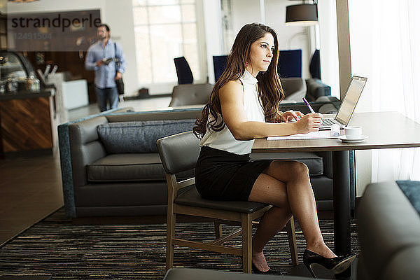 Eine junge  schöne Frau sitzt auf einem Stuhl und schreibt etwas in ein Notizbuch  das vor ihr auf einem Tisch liegt. Links von ihr steht ein Laptop  rechts eine Tasse Tee.
