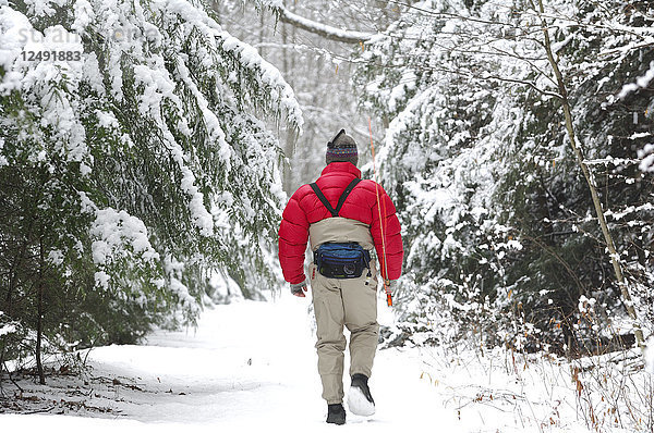 Ein Mann geht an einem kalten  verschneiten Wintertag mit seiner Angelausrüstung durch den Wald  um einen guten Platz zum Fliegenfischen zu finden.
