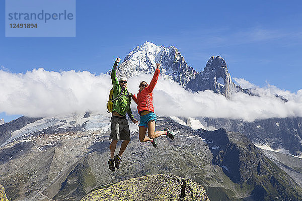Ein Mann und eine Frau in farbenfroher Kleidung springen fröhlich in die Luft  während im Hintergrund die schönen Berge von Chamonix in den französischen Alpen zu sehen sind.