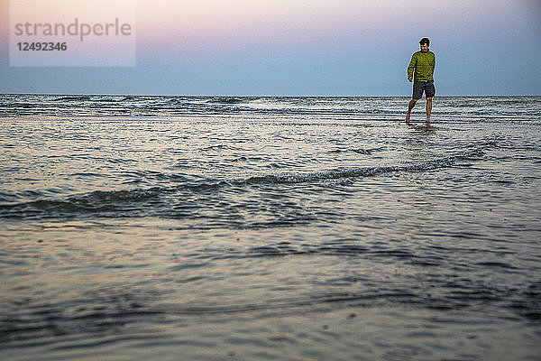 Ein Mann in Shorts und Jacke geht unter einem lavendelfarbenen Abendhimmel durch seichtes Wasser.