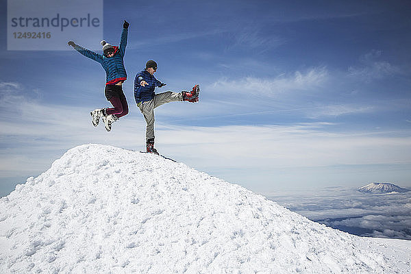 Eine Frau springt in die Luft und ein Mann kickt ein Bein hoch  um das Erreichen eines Berggipfels zu feiern.