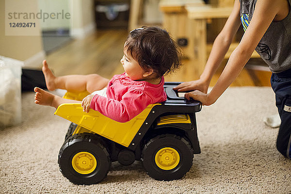 Ein einjähriges japanisch-amerikanisches Mädchen wird von ihrem älteren Bruder in einem Spielzeug-Kipplaster geschoben.
