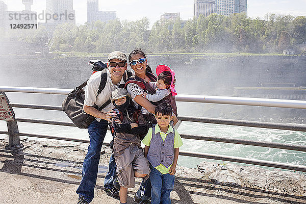 Eine Touristenfamilie  bestehend aus einem 6-jährigen japanisch-amerikanischen Jungen  einem 4-jährigen japanisch-amerikanischen Jungen  einer japanisch-amerikanischen Mutter und ihrem 6 Monate alten Baby sowie einem amerikanischen Vater  der die Niagarafälle von der Seite der Niagarafälle  New York  USA  betrachtet. Der Hintergrund ist ein Teil der Skyline von Niagara Falls  Ontario  Kanada.
