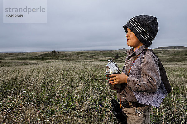 Ein 4 Jahre alter japanisch-amerikanischer Junge  der als Forscher mit Hut und Weste gekleidet ist  untersucht das Land (Grasland und Prärien) und hält seine alte Metallflasche im Badlands National Park  South Dakota.