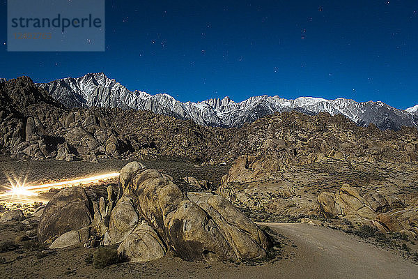 Autolichter streifen durch eine nächtliche Landschaft mit Geröllhalden und einer verschneiten Bergkette mit zerklüfteten Gipfeln in der Ferne.