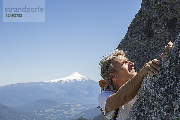 Porträt eines reifen Mannes  der auf einem Felsen mit einem Vulkan im Hintergrund klettert