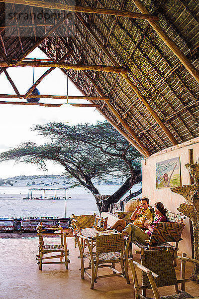 Ein Mann und eine Frau sitzen in einer strohgedeckten Sitzecke mit Blick auf einen afrikanischen Strand.