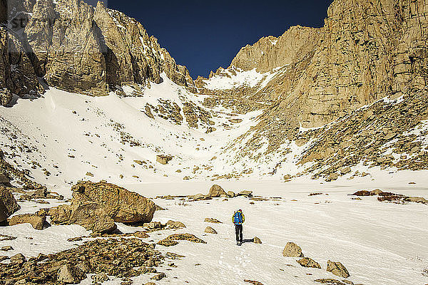 Ein 30-jähriger Mann in Bergsteigerkleidung geht ein schneegefülltes Tal mit zerklüfteten Granitwänden hinauf.