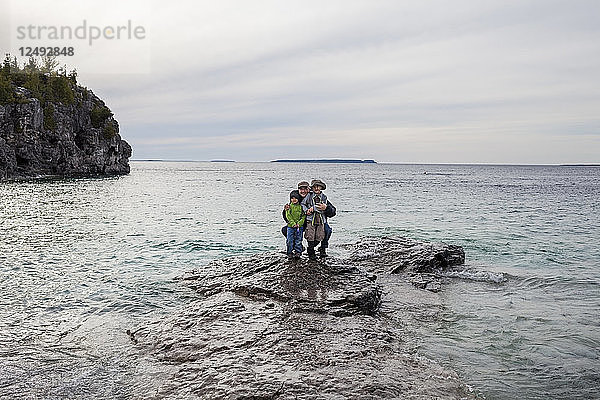 Eine Familie  bestehend aus einem 4-jährigen japanischen Amerikaner  einem 6-jährigen japanischen Amerikaner und einem Vater  steht auf einem Felsen im seichten Huronsee  während sie die Grotte im Bruce Peninsula National Park  Ontario  Kanada  erkunden.