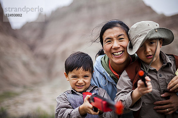 Eine japanisch-amerikanische Frau und Mutter hockt mit ihrem 4 Jahre alten japanisch-amerikanischen Jungen und ihrem 6 Jahre alten amerikanischen Jungen  die jeweils mit Spielzeugpistolen  einer 6-Schuss-Pistole und einem Gewehr ausgerüstet sind  inmitten von erodierenden Felsformationen (Zinnen und Türme) im Badlands National Park  South Dakota.