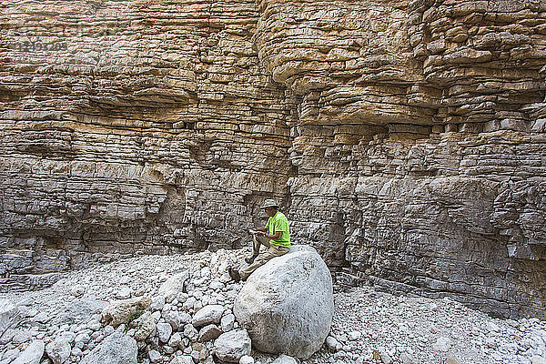 Älterer Mann sitzt auf einem Felsen in einer schmalen trockenen Schlucht mit steilen  geschichteten Felswänden