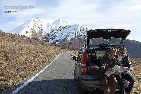 Ehepaar schaut vom Kofferraum eines Autos auf einer Bergstraße auf die Karte