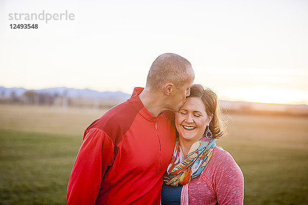 Ein Mann küsst seine lächelnde Frau auf den Kopf  während er auf einem Feld bei Sonnenuntergang steht