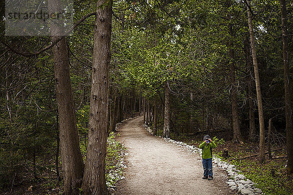 Ein 4 Jahre alter japanisch-amerikanischer Junge geht einen Waldweg entlang und macht ein Foto mit seiner Spielzeugkamera  während er den Bruce Peninsula National Park  Ontario  Kanada  erkundet.