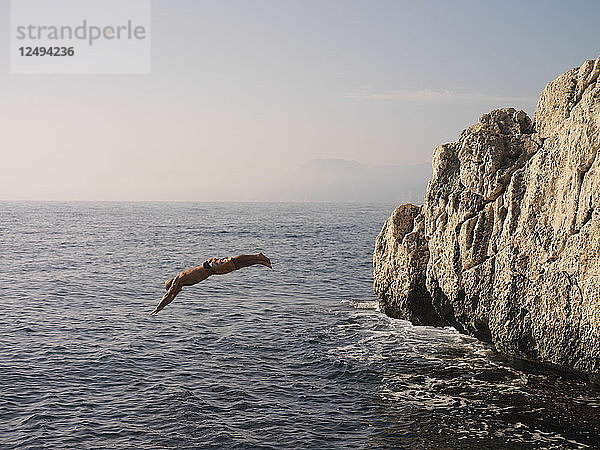 Mann springt bei Sonnenuntergang von einer Felswand ins Meer