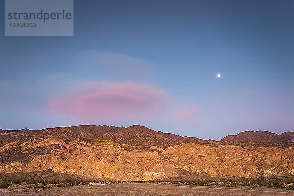 Der Mond geht an einem dunkelblauen Himmel mit einer rosafarbenen Wolke auf  die über einem Wüstengebirge hängt  das von der untergehenden Sonne orange beleuchtet wird.