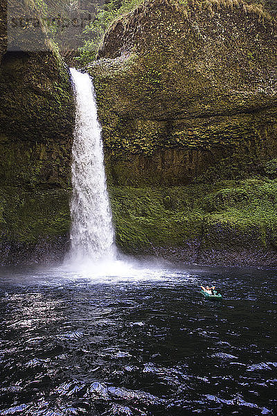 Ein Mann schwimmt in einem Ein-Personen-Rafting in einem dunklen Pool unter einem hohen Wasserfall.