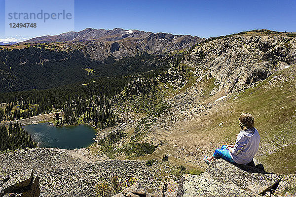 Eine junge Frau sitzt auf einem Felsvorsprung in den Bergen mit Blick auf einen See in Colorado