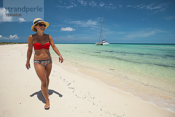 Eine junge Frau mit Bikini und Sonnenhut spaziert im Urlaub auf Kuba über einen Sandstrand.