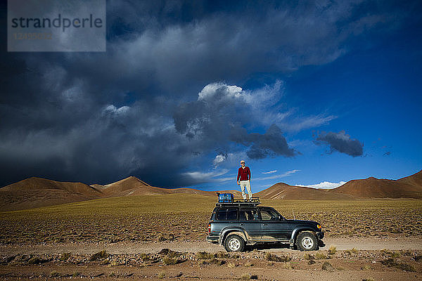 Ein Mann steht auf dem Dach seines Lastwagens  um ein Foto des dramatischen Himmels bei Sonnenuntergang in der Nähe des Salar de Uyuni in Bolivien zu machen.