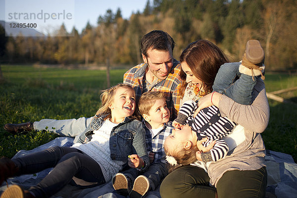 Eine fünfköpfige Familie lacht gemeinsam  während sie auf einer Decke im Freien sitzt.