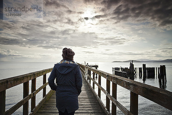 Eine Frau in einem blauen Mantel und einer Strickmütze geht auf das Ende des Davis Bay Piers in Secelt  B.C.  Kanada zu.