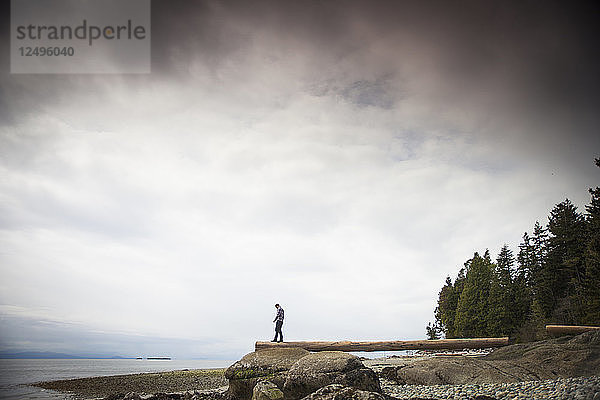 Ein junger Mann balanciert auf einem Baumstamm mit Blick auf den Pazifischen Ozean im Robert's Creek Park in der Nähe von Sechelt  BC  Kanada.