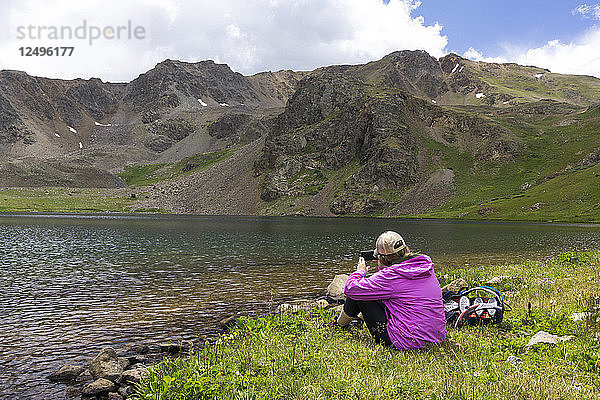 Eine junge Frau sitzt an einem hochalpinen See und macht ein Foto mit ihrem iPhone