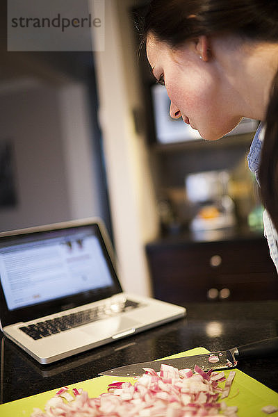 Eine junge Frau liest ein Rezept auf ihrem Laptop  während sie in der Küche kocht.