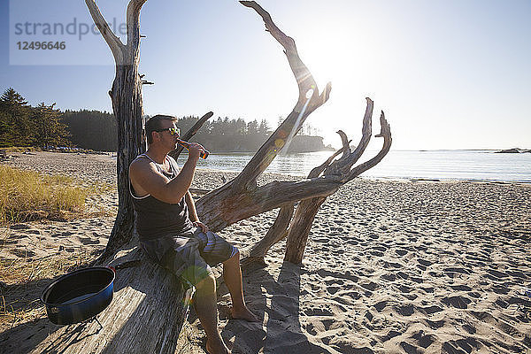 Ein Mann sitzt auf einem Baumstamm und trinkt Bier am Strand