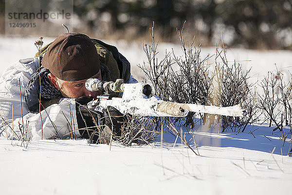 Ein Jäger in Kältetarnkleidung zielt mit einem Gewehr auf sein Ziel  während er sich in einem schneebedeckten Feld in der Nähe von Merritt  British Columbia  Kanada  hinlegt.