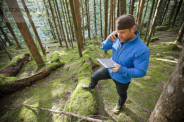 Ein Mann telefoniert  während er im Wald an einem Tablet arbeitet.